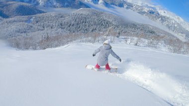 Unrecognizable active tourist snowboards in the scenic Slovenian backcountry on a sunny winter day. Athletic young woman snowboards in the breathtaking mountains of the idyllic white Julian Alps.