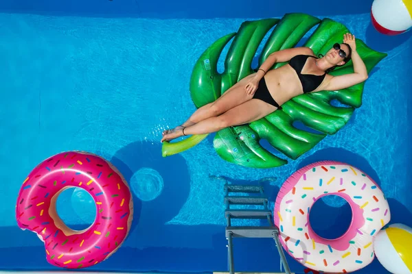 Caucasian female in a black bikini relaxes in her garden pool on a sunny summer weekend. Beautiful young woman lies on a big leaf floatie in her crystal clear home pool.