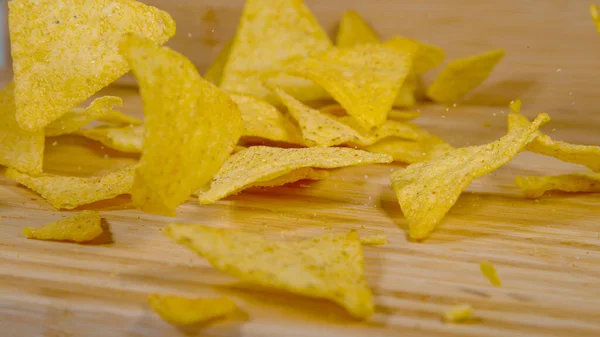 CLOSE UP, DOF: Delicious golden tortilla crisps fall on the wooden dining table and break into tiny bits and pieces. Unhealthy junk food falls on the empty wood kitchen countertop during a fun party.