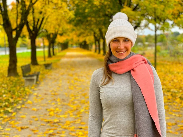 Happy female wears scarf and hat while going for a walk in park in fall. Cheerful Caucasian woman smiles while exploring the fall colored park on cold November day.