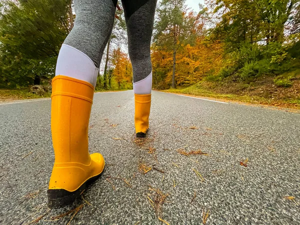 女性旅行者は風光明媚な森林ルートを歩いている間にゴム製のブーツを身に着けています 黄色のラバーブーツとグレーのヨガパンツを身に着けている女性は ぬれたアスファルトの道路に沿って歩く — ストック写真