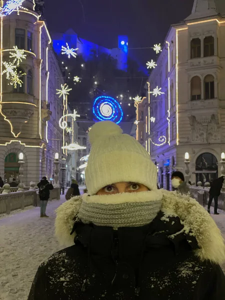 Verical Portrait Close 若い白人女性がリュブリャナのダウンタウンの明るいクリスマスライトを見ています お祝いの街の通りで帽子とスカーフを身に着けている女性観光客夜 — ストック写真