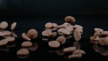 MACRO DOF: Bittersweet morsels of confection chocolate get scattered across a shiny black kitchen table. Cinematic shot of milk chocolate droplets falling onto a polished countertop. Falling chocolate