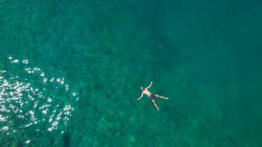 AERIAL, TOP DOWN: Flying above a relaxed Caucasian male tourist floating on his back during a swim in the turquoise colored ocean. Carefree bearded man swims on his back in the gorgeous emerald sea.