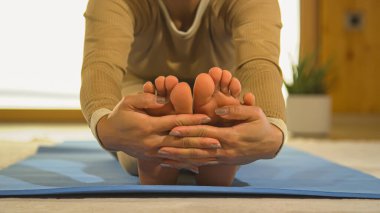 Front view of woman before leaning into seated forward fold yoga posture. Young female person doing body stretching on blue yoga mat. Healthy indoor activity for vitality and flexibility.
