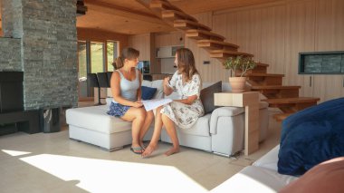 CLOSE UP: Two young women sitting on a sofa and consulting interior house design. Home owner and design expert discussing house building plans to efficiently organize and arrange home living space clipart