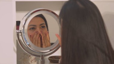 Young lady self massaging her pretty face in cosmetic mirror reflection. Beautiful woman during relaxing face treatment for facial muscle relief. Female person at facial beauty treatment.