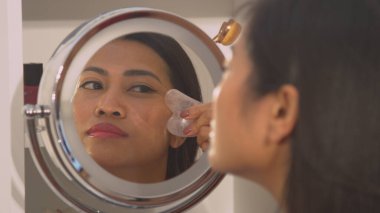 Young lady self massaging her face with jade scraper for facial lifting. Reflection of beautiful Asian woman in cosmetic mirror performing anti-aging massage treatment with gua sha scraper.