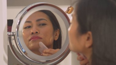 Reflection of beautiful woman in cosmetic mirror using gua sha scraper. Young Philippine lady at rejuvenating beauty treatment using jade roller for skin firmness and face lifting massage.