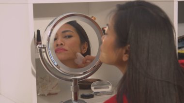 View of pretty young lady in mirror reflection using gua sha scraper. Young Philippine lady at rejuvenating beauty treatment using jade roller for skin firmness and face lifting massage.