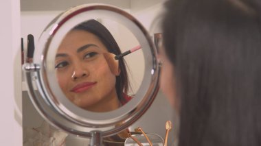Attractive Philippine woman applying bronzer to beautify her face. Mirror reflection of a pretty lady using brush to apply face blush. Female person taking care of her fresh look.