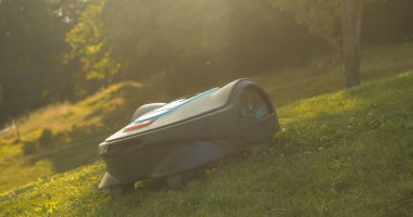 Autonomous lawn mower moving downhill and trimming grass in backyard. Lawn robot, backlit with golden sunlight, cutting green turf in modern garden. Futuristic gardening equipment at work.