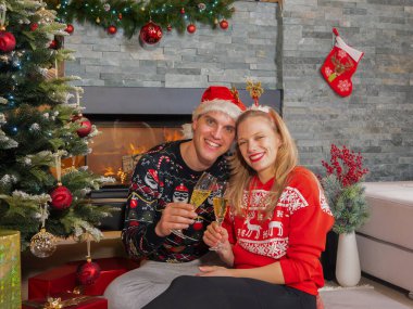Cute cheerful couple toasts with glasses of champagne on Christmas Eve. Happy young man and woman enjoying and celebrating with champagne sitting by Christmas tree in front of a fireplace.