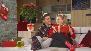 Joyful couple wearing Christmas sweaters and exchanging gifts on festive evening. Cheerful young man delighted his beautiful girlfriend by giving her a nicely wrapped present for Christmas holidays.