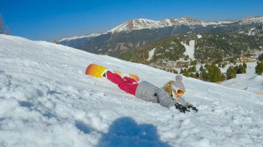 Young woman trying to learn to ride a snowboard when she crashes into soft snow. Female beginner snowboarder is sliding face forward after falling down on a snowy ski slope at winter ski resort.