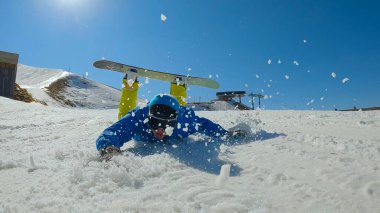 Man falls on snow and slides towards camera while trying to snowboard. Male snowboarder lands into snow after losing balance at snowboarding down the slope at snowy ski resort on sunny day.