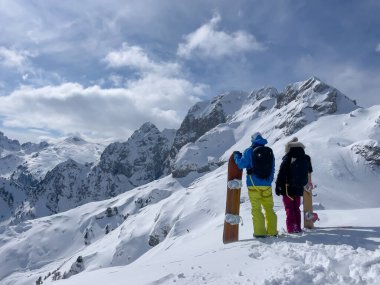 Freeride couple admiring endless possibilities for snowboarding fresh powder snow. Young woman and man holding snowboards on top of snowy mountain ridge. Amazing views in pristine Albanian mountains.