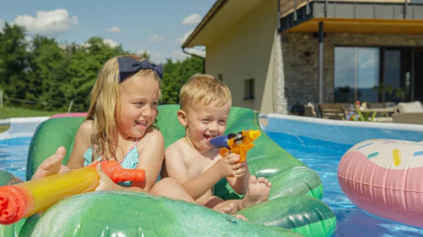 CLOSE UP: Two happy children smiling and having fun in the backyard swimming pool. Sister and brother enjoying at pool party floating on inflatable toys on a sunny summer day. Water games for hot days