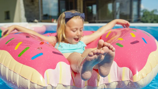 CLOSE UP: Cute girl floating in pool on inflatable donut and splashing with legs. Cheerful kid enjoying while having fun in home swimming pool. Outdoor summer activities for hot days in home backyard.