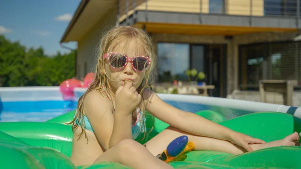 PORTRAIT: Sweet girl enjoying ice cream bar in the backyard pool on a hot day. Adorable girl eating chocolate ice cream bar in backyard garden pool. Complete refreshment on a hot and sunny summer day.