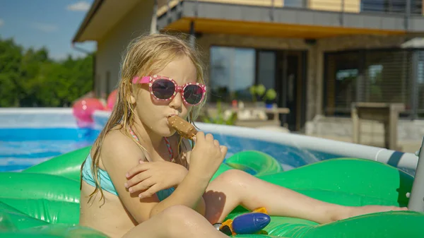 CLOSE UP: Adorable girl floating in pool and eating ice cream on a hot summer day. Cute little girl enjoying chocolate ice cream bar in backyard garden pool. Complete refreshment on a hot summer day.
