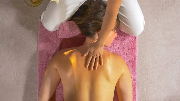 Therapeutic masseuse massaging upper back and shoulder of a young lady. Female massage therapist using massage oil for deep muscle relaxation. Wellness treatment for back muscle relief.