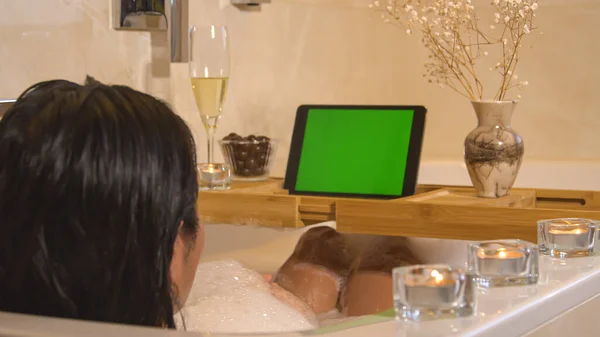Young lady relaxing and chilling in bubble bath while watching series. Beautiful woman relaxing at home bathroom and taking time for herself. Home wellness for stress relief and body care.
