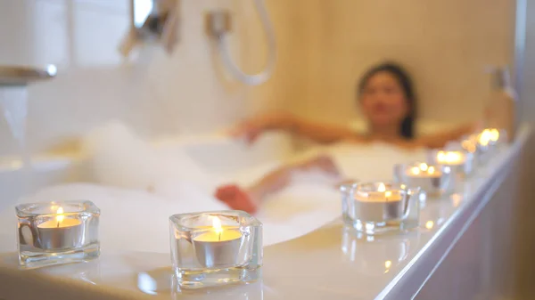 Lined up glowing candles for relaxing atmosphere while taking a nice bubble bath. Beautiful young Asian woman treating herself with spa and relaxing while chilling in nice bath full of bubbly foam.