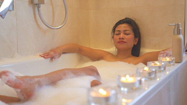 Расслабленная женщина с закрытыми глазами наслаждается пузырьковой ванной в окружении свечей. Подкладка светящиеся свечи для расслабляющей атмосферы и приятного опыта во время проведения спа-процедуры в домашней ванной комнате.
