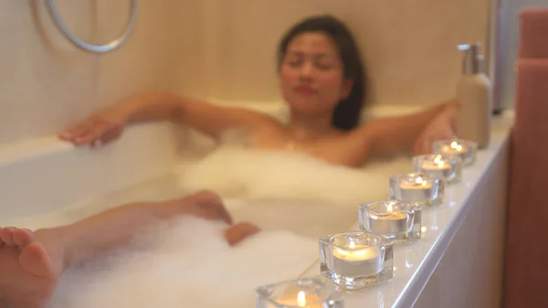 Attractive lady with eyes closed chilling in a bubbly bath embraced with candles. Young Asian woman relaxing in bathtub. Lined up glowing candles for relaxing atmosphere and pleasant spa experience.