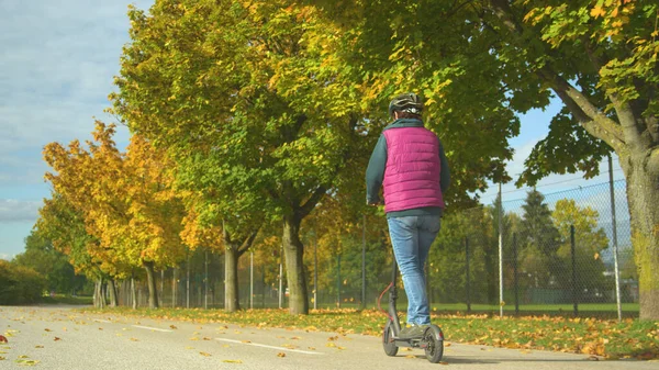 Rear view of an elderly woman driving an electric scooter next to autumn trees. Senior lady using modern end eco-friendly way of commuting. Beautiful autumn day to ride with e-scooter around the town.