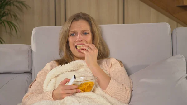 Young lady becoming upset while watching latest television news report. Pretty woman covered with blanket and eating snacks while following TV news. Woman on a comfy sofa watching TV report.