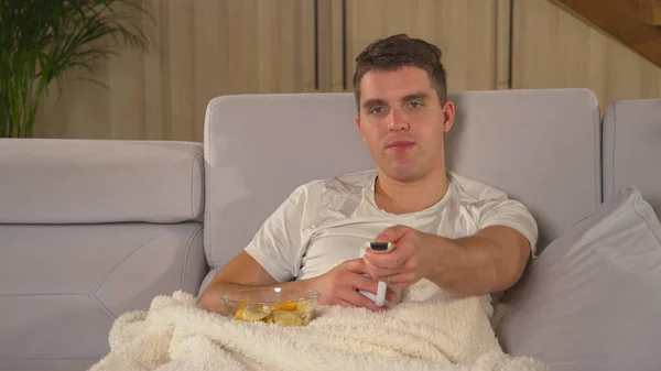 Man enjoying on a comfy couch, drinking tea and watching television. Young guy switching TV programs while chilling covered with blanket on sofa. Male person relaxing in home living room.