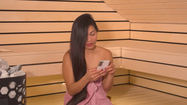 Pretty Filipina Wrapped Towel Sitting Bench Sauna Using Smartphone Attractive — 图库照片