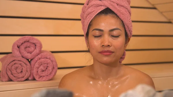 Face shot of Filipina with eyes closed sweating in hot Finnish sauna. Lady at sauna treatment for detoxification, stress relief and strengthening of the immune system in cold autumn months.