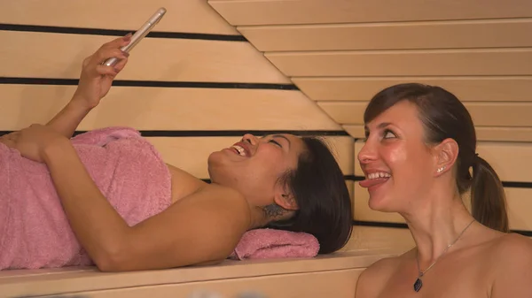 Smiling young women creating memories from relaxing at sauna and spa. Beautiful girlfriends wrapped in pink towels enjoying and having fun at wellness treatment in wooden Finnish sauna.