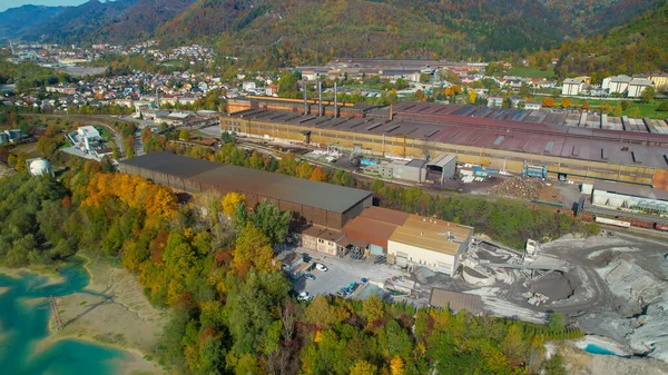 Industrial infrastructure near city and mountains in vibrant fall season. Steel factory located by the river in picturesque valley surrounded with alpine landscape in colorful autumn shades.