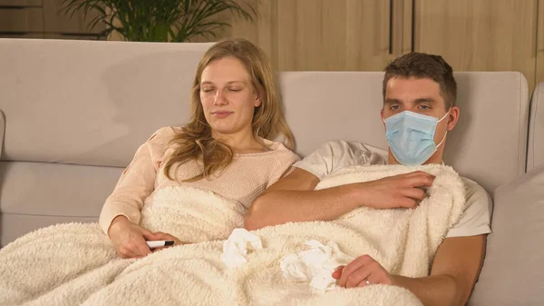 Young man with a face mask is careful not to spread virus to his lady. Young couple resting on comfy couch during covid quarantine. Increase of diseases and virus spread in autumn time.