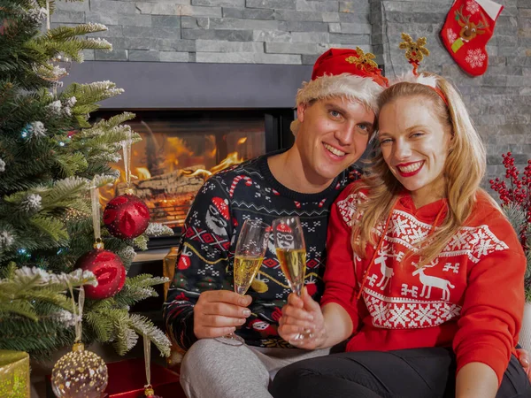 Smiling young couple toasts with glasses of champagne on New Years Eve. Happy young man and woman enjoying and celebrating with champagne sitting by Christmas tree in front of a fireplace.