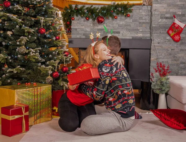 かなり若い女性は 贈り物で彼女を驚かせ 喜んだ男を抱きかかえています 素敵なカップルは クリスマス休暇を祝い 暖炉の前で美しく装飾された木のプレゼントを交換します — ストック写真