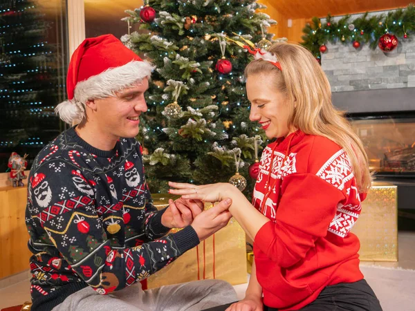 愛の幸せなカップルは お祝いの夜にクリスマスツリーの横に従事取得します 若い男は素晴らしいロマンチックなジェスチャーで彼の女性を驚かせ クリスマスに婚約指輪で彼女に提案します — ストック写真