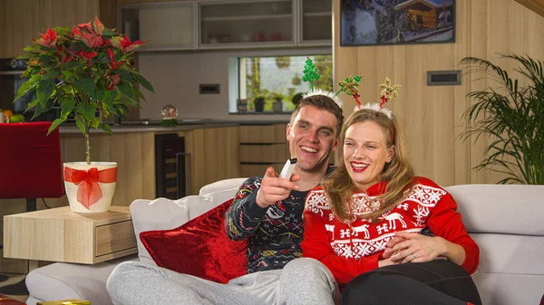 Lovely young couple relaxing on comfy couch and watching Christmas comedy on TV. Man and woman sitting on cosy sofa in home living room and enjoying at funny holiday television entertainment program.