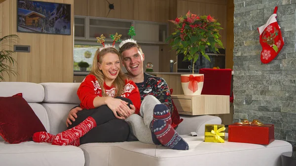Christmas couple enjoying and laughing while watching amusing television program. Cheerful man and woman sitting on comfy couch in decorated living room and relaxing at funny Christmas movie comedy.