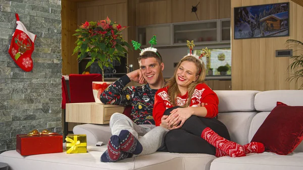 Cute couple in Christmas outfit laughing while watching holiday movie comedy. Cheerful man and woman sitting on comfy couch in decorated living room and relaxing at amusing TV entertainment program.