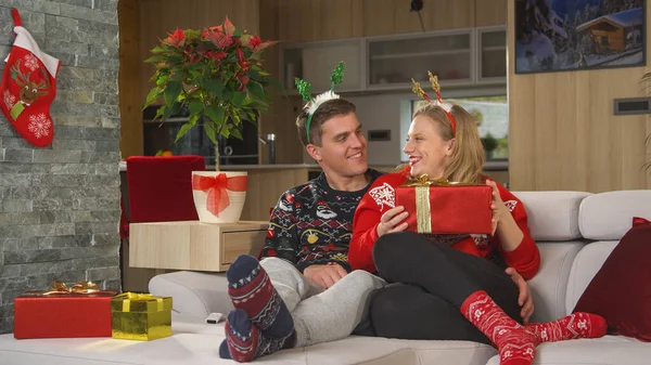Joyful couple wearing Christmas sweaters and exchanging gifts on festive evening. Cheerful young man delighted his beautiful girlfriend by giving her a nicely wrapped present for Christmas holidays.
