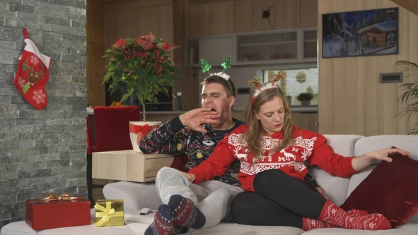 Young man yawning and getting tired while watching TV program on Christmas Eve. Young couple in Christmas sweaters relax together on comfy sofa in home living room after celebrating festive holidays.