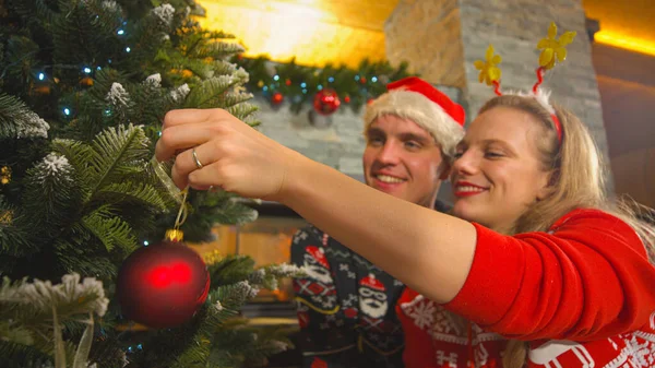 美しい女性は彼女の男と一緒にクリスマスツリーに赤ん坊を吊るす 素敵な若いカップルは お祝いの服を着て 美しく装飾された家のリビングルームでクリスマス休暇を祝います — ストック写真