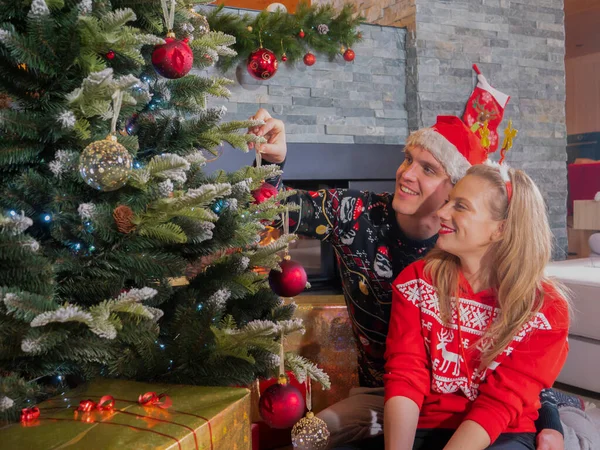 暖炉に座っているロマンチックなカップルとバーベキューでクリスマスツリーを飾る お祝いの冬の休日を祝うために自宅のリビングルームでクリスマスの装飾を準備する若い男と女 — ストック写真