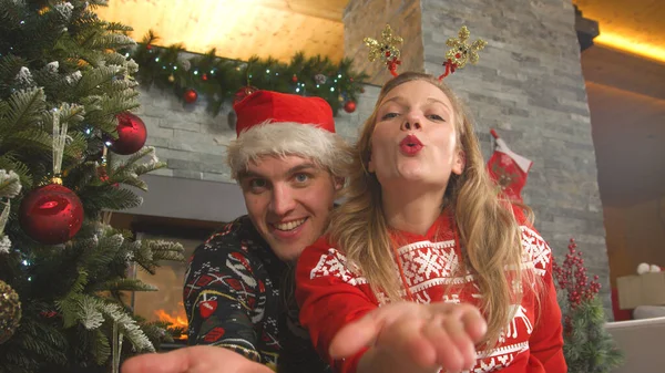 カメラに向かってキスを吹いているクリスマスのセーターの楽しい二人組 笑顔の男と女性はクリスマスのイブを祝いながら陽気な感じ 若いカップルポーズ装飾木の横にある — ストック写真