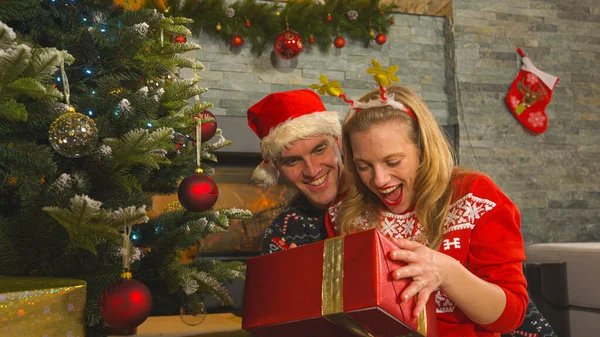 若い女性は夫から贈られたクリスマスプレゼントに喜んでいます 飾り木の下でクリスマスプレゼント 暖かい家の抱擁でお祝い12月の休日を祝うかわいいカップル — ストック写真